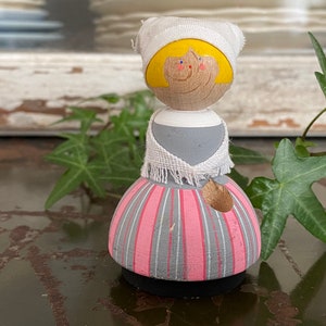 May include: A wooden doll with a pink and gray striped skirt, a white shawl, and a white cap with yellow hair. The doll has a painted face with yellow eyes and a smile.