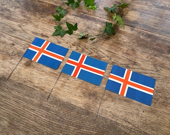 May include: Three small Icelandic flags with blue backgrounds and a white-bordered red cross, are displayed on thin metal sticks. The flags are arranged on a wooden surface, with a green vine in the background.