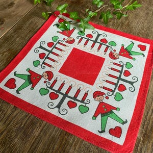 May include: Square white and red Christmas-themed cloth with a red border. The cloth features a central red square, surrounded by illustrations of candle holders, elves, hearts, and candles. The design is festive.