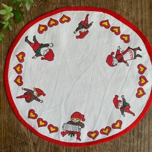 May include: A white round tablecloth with a red border and a repeating pattern of red hearts with yellow centers. The tablecloth features illustrations of seven cartoon figures wearing red hats and playing musical instruments.