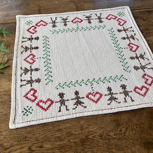 Pode incluir: Um pano quadrado bordado em creme com uma borda de figuras marrons de mãos dadas, corações vermelhos e detalhes verdes. O desenho está centrado em uma superfície de madeira.