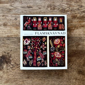 Könnte beinhalten: Ein Buch mit dem Titel "FLAMSKVÄVNAD" mit einem weißen Einband mit farbenfroher Textilkunst. Das Cover zeigt stilisierte Figuren, Vögel und florale Designs in Rot, Schwarz und Gold. Das Buch liegt auf einer Holzoberfläche.