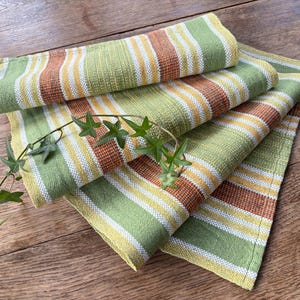 May include: Four folded green, yellow, white, and brown striped cloth napkins are stacked on a wooden surface. A green vine with small leaves is draped across the napkins. The napkins have a woven texture.