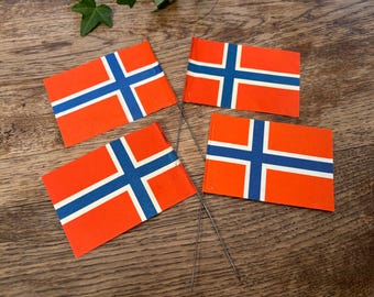 May include: Four small Norwegian flags with red backgrounds, blue crosses, and white borders are arranged on a wooden surface. The flags are rectangular and attached to thin metal sticks, suggesting they are decorative or for display.