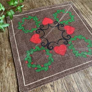 May include: A brown linen placemat with a white border features a repeating pattern of green wreaths with red hearts in the center. The wreaths are connected by black swirls.