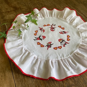 May include: A round, white placemat with a red ruffled edge. The center features a ring of cartoon figures in red and black outfits, interspersed with red and yellow hearts. The placemat is on a wooden surface.