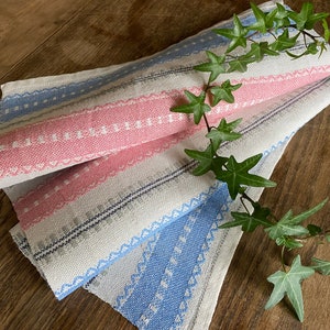 May include: Two woven kitchen towels with pink and blue stripes and white accents. The towels are folded and layered on top of each other. A green vine with leaves is draped over the towels.