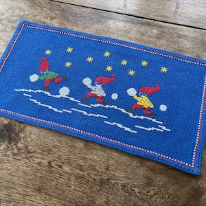 May include: A blue rectangular needlepoint featuring three figures in red, green, and yellow outfits, playing in the snow. The figures are under yellow star-shaped designs. The border is red and white.