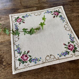 Pode incluir: Toalha de mesa quadrada creme com uma borda floral com rosas cor-de-rosa e flores azuis. Uma videira verde com folhas é drapeada sobre a toalha de mesa. A toalha de mesa está sobre uma superfície de madeira.