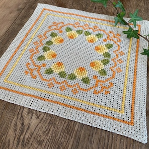May include: A white crocheted square placemat with a border of orange and yellow thread. The center features a circular design of yellow flowers with green leaves.