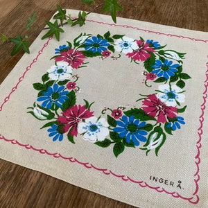 May include: A white linen table runner with a pink border and a floral wreath design. The wreath is made up of pink, blue, and white flowers with green leaves. The text "INGER A." is printed in the bottom right corner.