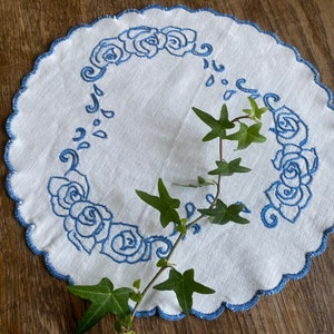 May include: A white crocheted doily with a blue embroidered floral design. The doily has a scalloped edge and is decorated with a repeating pattern of roses and leaves.