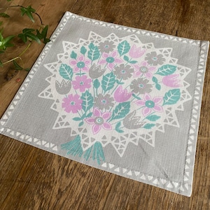May include: A gray linen placemat with a white floral design featuring pink and green flowers. The design is centered on the placemat and surrounded by a white lace border.