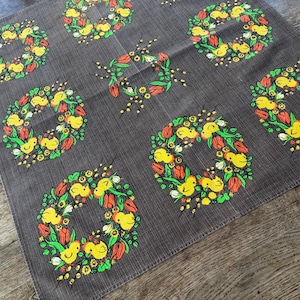 Könnte beinhalten: Ein dunkelgrauer quadratischer Stoff mit sich wiederholenden kreisförmigen Kranzdesigns. Jeder Kranz besteht aus gelben Küken, roten Tulpen, grünen Blättern und gelben Blumen. Der Stoff ist wahrscheinlich ein dekoratives Textil.