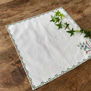 May include: White linen napkin with a green and brown embroidered border and a small floral design in the corner. The napkin is on a wooden surface with a green vine.