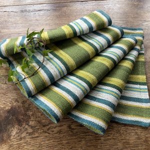 May include: Folded green, white, and blue striped fabric napkins. The napkins are arranged in a fan shape, with a sprig of green leaves resting on top. The fabric has a textured appearance, and the stripes are of varying widths.