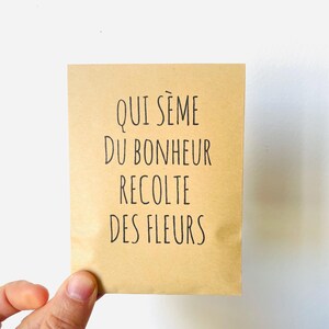 Peut inclure: Une petite carte rectangulaire avec le texte français "QUI SÈME DU BONHEUR RECOLTE DES FLEURS" en lettres noires. La carte est tenue sur un fond blanc. La carte est de couleur marron clair.