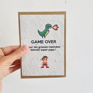 Può includere: Un biglietto di auguri con un drago verde pixelato che sputa fuoco e un personaggio pixelato in rosso. Il biglietto recita "GAME OVER" e "our tes grasses matinées bientôt super papa!" su uno sfondo bianco strutturato, incorniciato da cartoncino marrone.