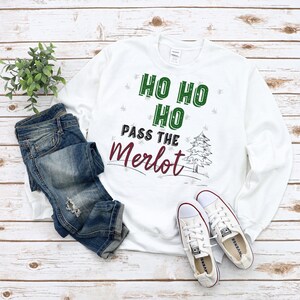 Puede incluir: Una sudadera blanca con el texto "HO HO HO PASS THE Merlot" en verde y burdeos. La sudadera se combina con vaqueros azules y zapatillas blancas, creando un atuendo casual y festivo. Una pequeña planta verde es visible en el fondo.