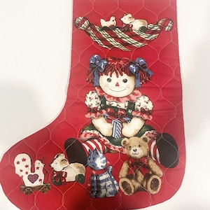 Peut inclure: Une chaussette de Noël rouge matelassée ornée d'une illustration centrale représentant une poupée aux cheveux en laine rouge avec des nœuds bleus, entourée d'un ours en peluche, d'un chat bleu, d'un oiseau blanc et d'une souris blanche.