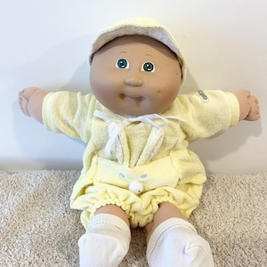May include: A doll with a round face, green eyes, and a dimple on the left cheek. It wears a yellow terry cloth outfit with a white ribbon tie, a matching yellow hat with white polka dots, and plain white socks on its feet. The doll has a tan complexion.