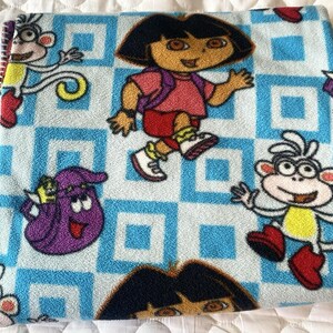 May include: A fleece blanket featuring a repeating pattern of Dora the Explorer, Boots the monkey, and Backpack against a light blue and white checkered background. The characters are depicted in their signature colours, including pink, purple, and yellow.