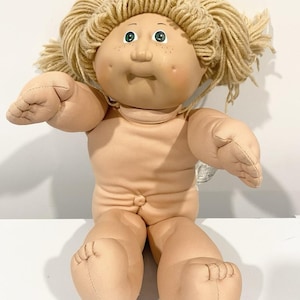 May include: A doll with a soft fabric body, light skin tone, and dimpled cheeks. It features yarn hair in a light blonde shade styled in pigtails, green eyes, and freckles. The doll has a stitched navel and is posed with arms extended forward.