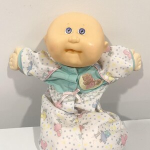 May include: A bald doll with a round face, light skin, and blue eyes. The doll wears a white sleepsuit with a pattern of small blue dots and colourful sheep illustrations, featuring a teal chest panel, a small pink bow, and a teddy bear patch.