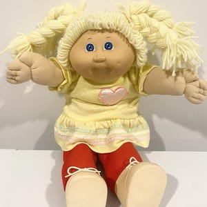 Peut inclure: Une poupée à la peau claire, aux yeux bleus et aux cheveux en laine blonde coiffés en deux nattes. La poupée porte un haut jaune à manches courtes avec un cœur rose appliqué, une jupe jaune à rayures pastel, un pantalon rouge et des chaussures beiges.