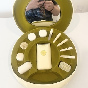 Peut inclure: Un coffret de soin Philips rond de couleur crème avec un intérieur vert olive. Le boîtier comprend un miroir intégré, un rasoir électrique central et divers accessoires de soin blancs disposés dans des compartiments moulés.