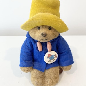 May include: A fuzzy brown bear figurine wearing a bright yellow hat and a blue duffle coat with two light-coloured toggles. A round white pin featuring a small illustration of a bear carrying a suitcase is attached to the front of the blue coat.