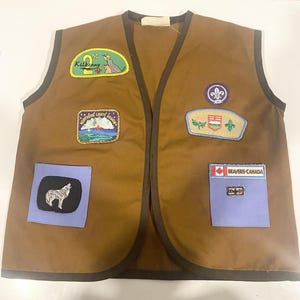 May include: A brown sleeveless waistcoat with dark brown trim featuring various embroidered patches, including 2nd Kilkenny, Northern Lights Region, Alberta Council Scouts Canada, and a Beavers Canada patch with a wolf graphic and a small rectangular label.