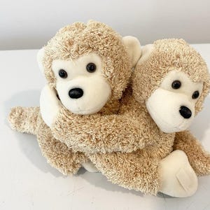 May include: Two plush monkey toys with textured tan fur and smooth cream-coloured faces. The monkeys have dark brown plastic eyes and black noses. One monkey is positioned behind the other, with its arms wrapped around the front monkey.