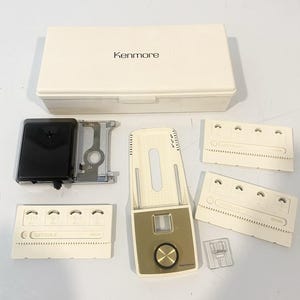 May include: A Kenmore sewing machine buttonhole attachment set including a rectangular off-white storage case, a black and silver metal mechanism, a central beige and gold control plate with a dial, three white plastic templates, and a clear presser foot.