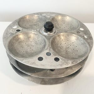Peut inclure: Un plateau à idli circulaire en aluminium doté de quatre moules concaves perforés pour la cuisson à la vapeur. Le plateau possède un bouton central noir pour la manipulation et comprend plusieurs petits trous de ventilation circulaires sur le pourtour.
