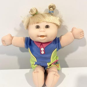 May include: A Cabbage Patch Kids doll with blonde hair styled in two top knots, brown eyes, and a light skin tone. The doll wears a blue and lime green collared outfit with two white buttons on the chest and a small graphic on the lower side.