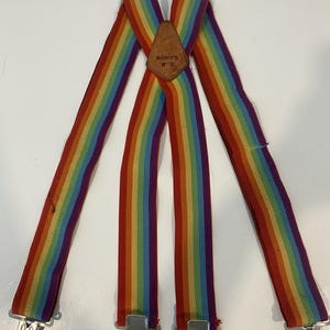 Peut inclure: Bretelles à rayures arc-en-ciel avec une pièce dorsale en cuir marron portant l'inscription KUNY'S SP-15. Les sangles élastiques présentent des bandes verticales rouges, orange, jaunes, vertes, bleues et violettes, avec quatre pinces métalliques.