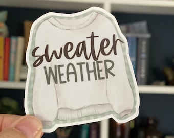 May include: White sweater sticker with the text "sweater weather" in brown and green lettering. The sticker has a green and white checkered border.