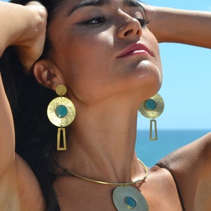 Peut inclure: Ensemble de bijoux dorés comprenant un collier et des boucles d'oreilles. Le collier est orné d'un grand pendentif circulaire avec une pierre turquoise. Les boucles d'oreilles ont un design similaire, avec un dessus circulaire et une forme rectangulaire. Le bijou est porté sur fond de ciel bleu et d'océan.