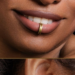 Puede incluir: Primer plano de los labios de una persona con un aro labial dorado. La imagen también muestra un brazalete de oreja dorado en la oreja. La joyería tiene un diseño simple y elegante. El tono de piel es oscuro y el fondo es negro.