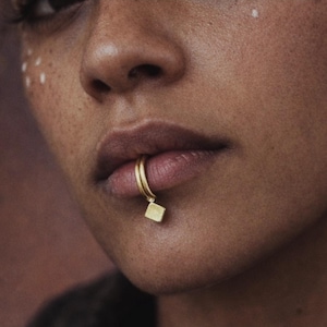 Puede incluir: Primer plano de un aro de labio dorado con un pequeño dije cuadrado. El anillo tiene un diseño de doble banda y el dije cuelga debajo del labio. El tono de piel es cálido con pecas.