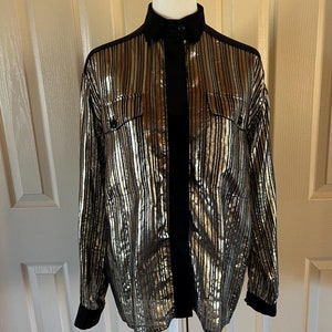 Puede incluir: Una camisa abotonada a rayas doradas y negras. La camisa tiene cuello, puños y una franja negra vertical en la parte delantera. Las rayas doradas son verticales y recorren toda la longitud de la camisa. La camisa se exhibe en un maniquí blanco.