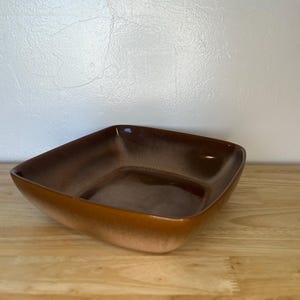 May include: A square, brown ceramic bowl with a glossy finish. The bowl has a gradient colour, transitioning from a lighter brown at the base to a darker brown at the rim. The bowl is empty and sits on a wooden surface.