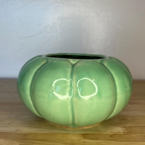 May include: A light green ceramic bowl with a pumpkin-like design. The bowl has a round shape with vertical ridges and a dark interior. It sits on a wooden surface.