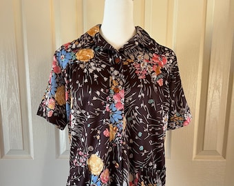 May include: A brown, short-sleeved button-down shirt with a floral pattern in shades of pink, yellow, blue, and white. The shirt has a collared neckline and two small pockets. The shirt is displayed on a mannequin.
