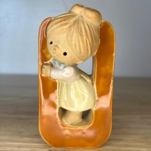 May include: A vintage ceramic figurine of a girl in a yellow dress and a blue bow, set within an orange-brown frame. The figurine has a classic, nostalgic design, with a detailed hairstyle and facial features. The overall aesthetic is reminiscent of mid-century decor.