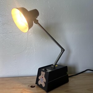 May include: A black desk lamp with a pivoting arm and a round, light-emitting shade. The base features a sticker with a teddy bear graphic and text. The lamp is plugged into an electrical outlet.