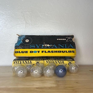 Puede incluir: Lámparas de flash Sylvania Blue Dot vintage en sus cajas originales. Las cajas son amarillas y negras con detalles en azul. Se ven varias bombillas de flash de cristal transparente, con una bombilla azul. Las cajas tienen la inscripción "SYLVANIA" y "BLUE DOT FLASHBULBS".