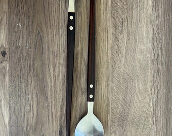 May include: A serving spoon and fork set. The spoon has a large, rounded stainless steel head and a dark wooden handle with two white dots. The fork has a similar design, with a slotted head and a matching handle.