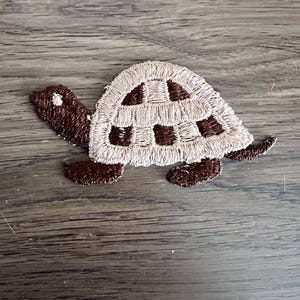 May include: A brown and beige embroidered turtle applique patch. The turtle has a checkered shell and brown legs.